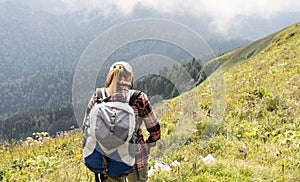Rear view of young woman in cap and plaid shirt putting on backpack hiking in green mountains in summer healthy active lifestyle,
