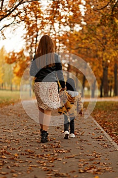 Rear view on a young woman in a fashion dress and a child in stylish clothes in the park. Mom and baby walks in the autumn park on