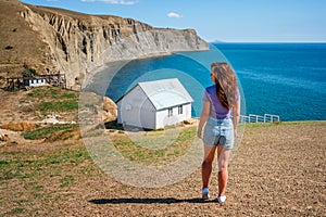 Rear view a young woman in front of a white lonely house on the edge of a cliff with a picturesque mountain landscape and a view