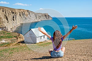 Rear view a young woman in front of a white lonely house on the edge of a cliff with a picturesque mountain landscape and a view