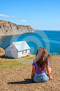 Rear view a young woman in front of a white lonely house on the edge of a cliff with a picturesque mountain landscape and a view