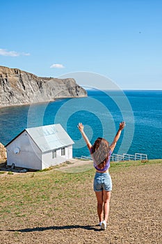 Rear view a young woman in front of a white lonely house on the edge of a cliff with a picturesque mountain landscape and a view