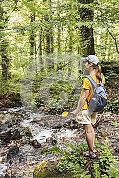 Rear view of young woman hiker in cap with backpack standing on mossy stone near mountain river in forest holding yellow fallen