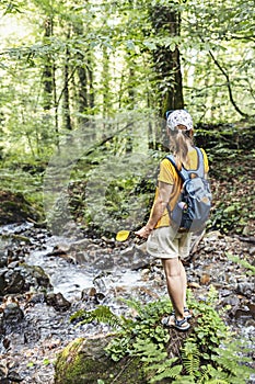 Rear view of young woman hiker in cap with backpack standing on mossy stone near mountain river in forest holding yellow fallen