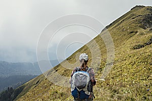 Rear view of young woman hiker in cap with large hiking backpack looking at the mountain view of the Aibga ridge of the Caucasus