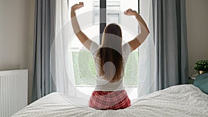 Rear view of young woman in pajamas sitting on the bed edge and stretching while looking out of the big window