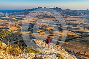 Rear view of young woman standing on mountain cliff and admiring beautiful landscape of coastal valley and sea. Teen tourist girl