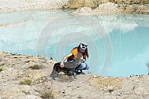 Rear view of young woman traveler in cap and ginger t-shirt stroking fluffy gray dog on sandy shore of blue lake or river, pet