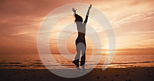 Rear view of young woman winner at sunset, jumping on the beach on sea background, raising her hands in the air, happy