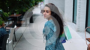 Rear of young female in dress in polka dot walking on street with packets and turning her head to camera. Beautiful