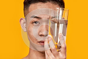 Rebalance. Portrait of young asian man looking at camera, holding a glass of water near his face isolated over yellow