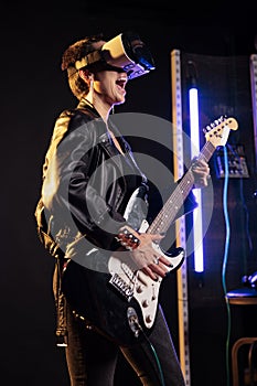 Rebel performer playing heavy metal song using eletric guitar while wearing vr headset to have rock concert simultation