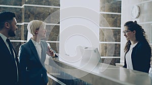 Receptionist girl talking with arrived businessman and businesswoman guests about check-in in hotel and giving key card