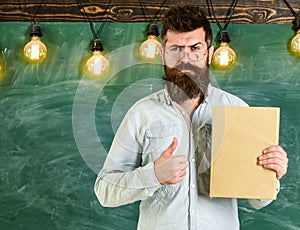 Recommendation concept. Bearded hipster holds book, chalkboard on background. Teacher in eyeglasses recommends book