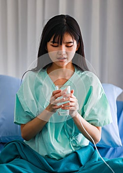 Portrait of recovered young asian woman patient sitting on bed in the hospital holding, looking to a glass of water wanting to