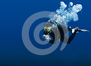 Recreational scuba diver with yellow mask, cloud of bubbles in black neoprene suit taking underwater photo in deep blue water of R