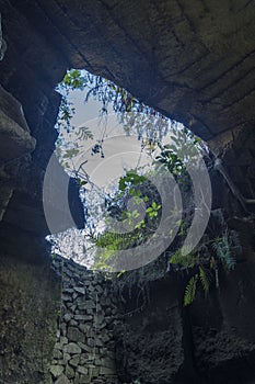 A rectangular opening in a dark, mossy limestone cave frames a bright blue sky with fluffy white clouds in Setigi