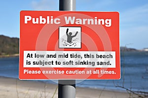 Red rectangular public warning sign with blurred beach background. Warning of soft sinking sand a low and mid tide.