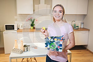 Recycling. Young smiling woman holding plastic lids in the container with green recycle icon on kitchen background