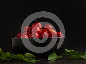 Red acerola cherries fruit in a ceramic bowl with a black background.