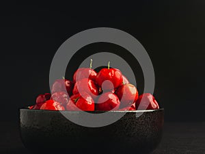 Red acerola cherries fruit in a ceramic bowl with a black background.