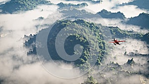Red airplane flying over mountains with pine trees in the clouds.