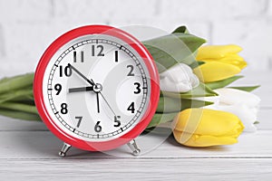 Red alarm clock and beautiful tulips on white wooden table against brick wall, closeup. Spring time