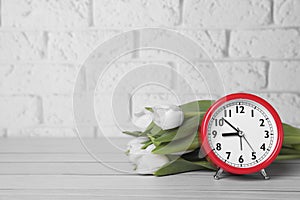 Red alarm clock and beautiful tulips on white wooden table against brick wall, space for text. Spring time