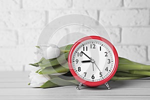 Red alarm clock and beautiful tulips on white wooden table against brick wall. Spring time