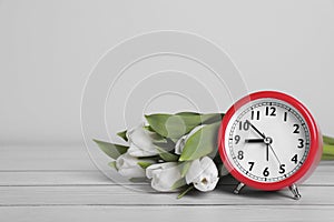 Red alarm clock and beautiful tulips on white wooden table against light background, space for text. Spring time