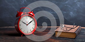 Red alarm clock, old books and reading glasses on a wooden table, black board background. 3d illustration