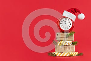 Red alarm clock in Santa Claus hat on wrapped gift boxes decorated fir branches and gold cones. Red background. Concept