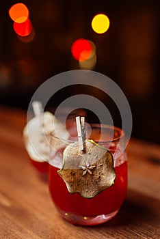 Red alcoholic cocktail decorated with dried pear slice on a wooden table in a bar. Blurry lights in the background.