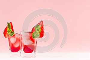 Red alcoholic shots of sweet fruit liquor with ice cubes, strawberry slice, green mint on pastel soft light pink background.