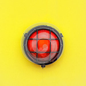 Red alert bulkhead light fixed to a painted yellow orange colour wall background. Photo in square format photo
