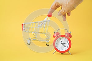 Red analog clock with blurred human`s hand and shopping cart on yellow background , sale promotion, payday concept