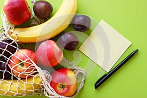 Red apples, bananas, plums, string bag and blank paper with black pen on a bright green background.