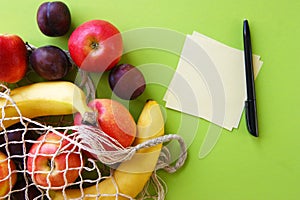 Red apples, bananas, plums, string bag and blank paper with black pen on a bright green background.