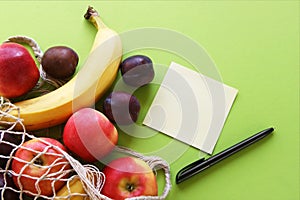 Red apples, bananas, plums, string bag and blank paper with black pen on a bright green background.