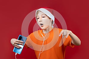 On a red background, a child with a Christmas hat and a yellow T-shirt holds a phone in his hand and listens to music through