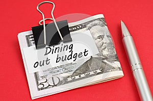 On a red background lies a pen and dollars clamped with a clip with the inscription on paper - Dining out budget