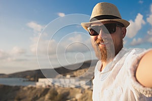 A red bearded hipster traveler in sunglasses and hat makes selfie on vacation on the seashore background