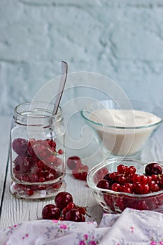 Red berries in a glass transparent bowl and sugar; currants in a jar on a light background