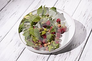 Red berries of wild raspberries in a white plate on a wooden table, a branch of raspberries with green leaves, organic
