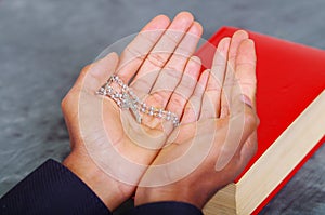 Red bible lying on blue desk with mans open hands above book, rosary cross placed inside, religion concept