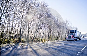 Red big rig American bonnet semi truck with dry van black semi trailer running on winter road with frosty trees