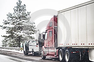 Red big rig broken semi truck with open hood and repair road assistant vehicle standing on the snowy winter road side shoulder