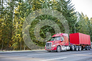 Red big rig low cab semi truck transporting container on the road with green forest on the side