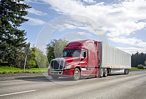 Red big rig popular bonnet semi truck transporting commercial cargo in dry van semi trailer moving on the straight wide highway