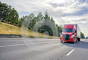 Red big rig semi truck with black grille transporting cargo in semi trailer driving on the straight wide highway road with trees